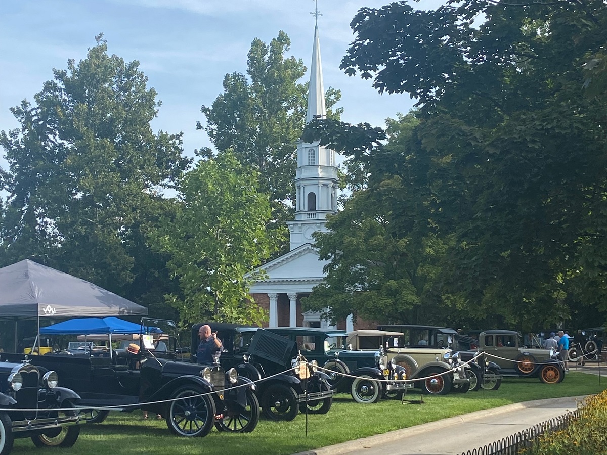 A row of cars are parked behind a string barrier on a lawn in front of a building with a white wood and red brick steeple; people stand by some of the cars