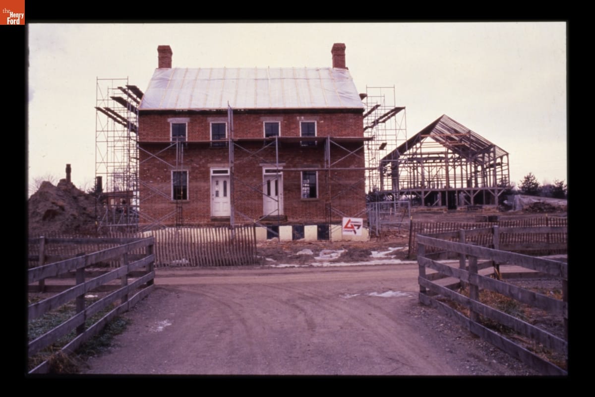 Firestone Farmhouse and Barn during Construction in Greenfield Village, December 1984 A two-story red brick house under construction, surrounded by scaffolding, with another timber-framed structure being constructed behind it