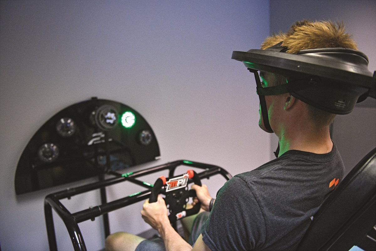 PitFit’s Race Trainer Man wearing large oblong headpiece sits holding what looks like a video game controller in front of a digital dashboard