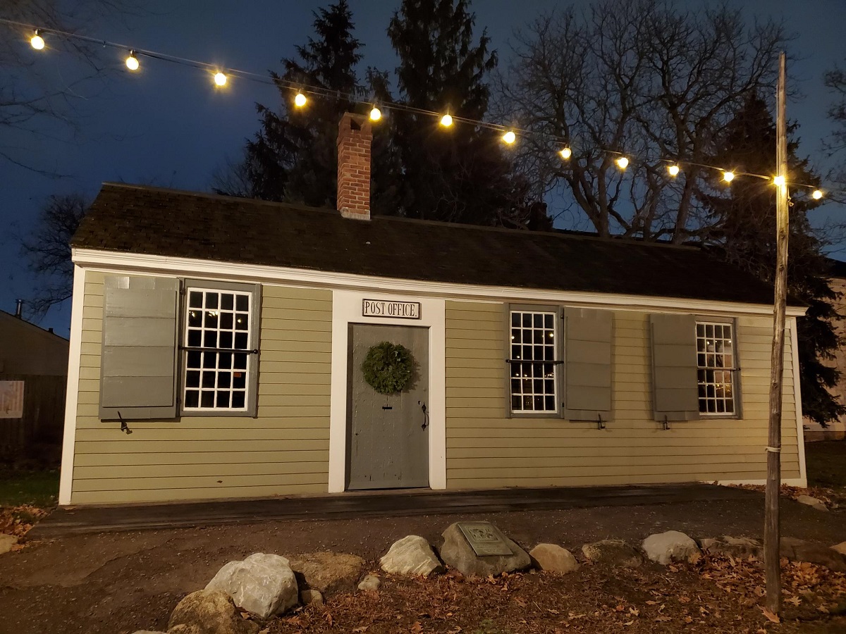 Phoenixville Post Office in Greenfield Village during Holiday Nights Small, beige, one-story wooden building with wreath on door and lights strung above it