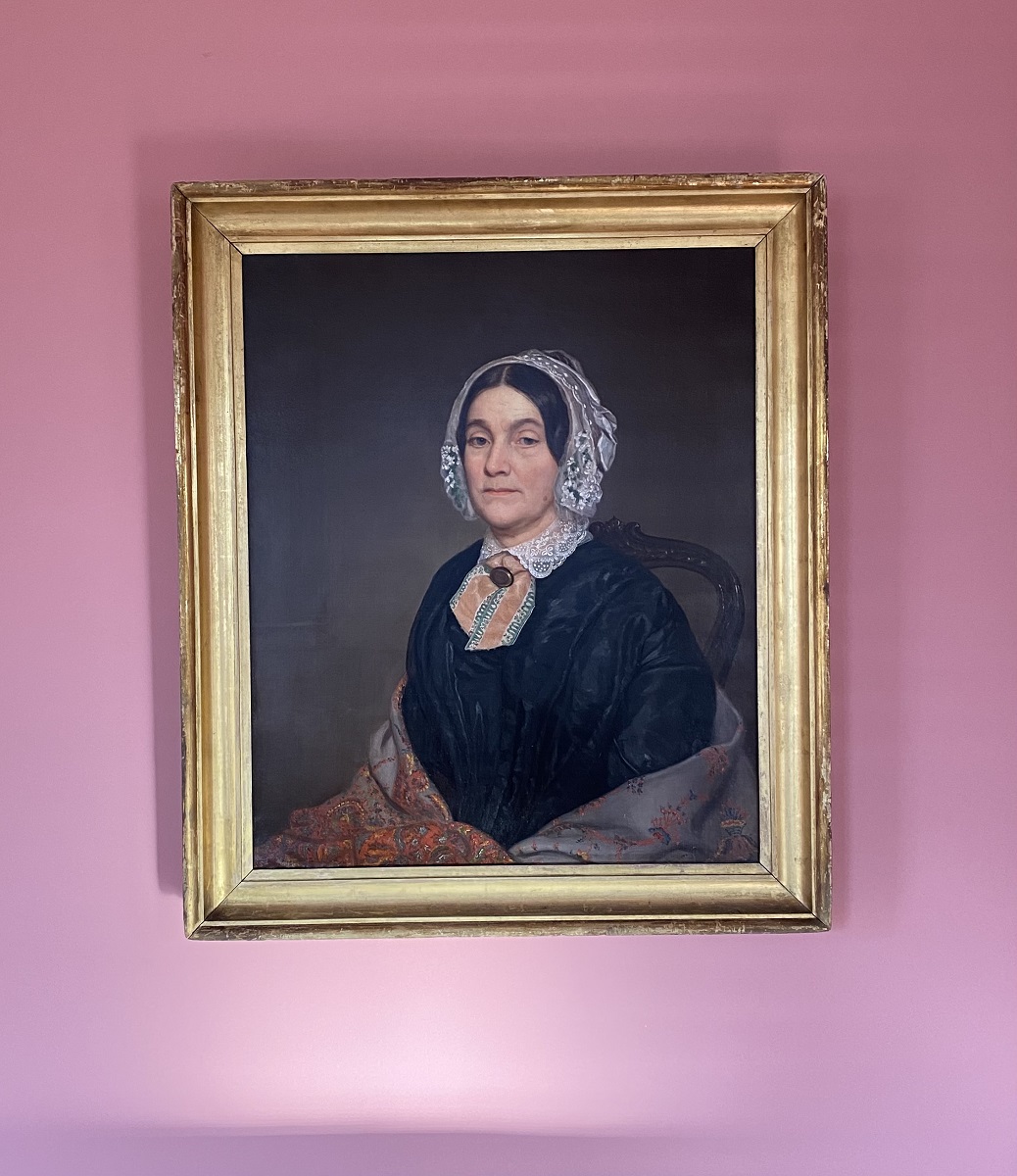 Treated “Lady in a Lace Bonnet” painting in Robert Frost Home Gold-framed portrait of woman in black dress, shawl, and white lace bonnet, hanging on pink wall