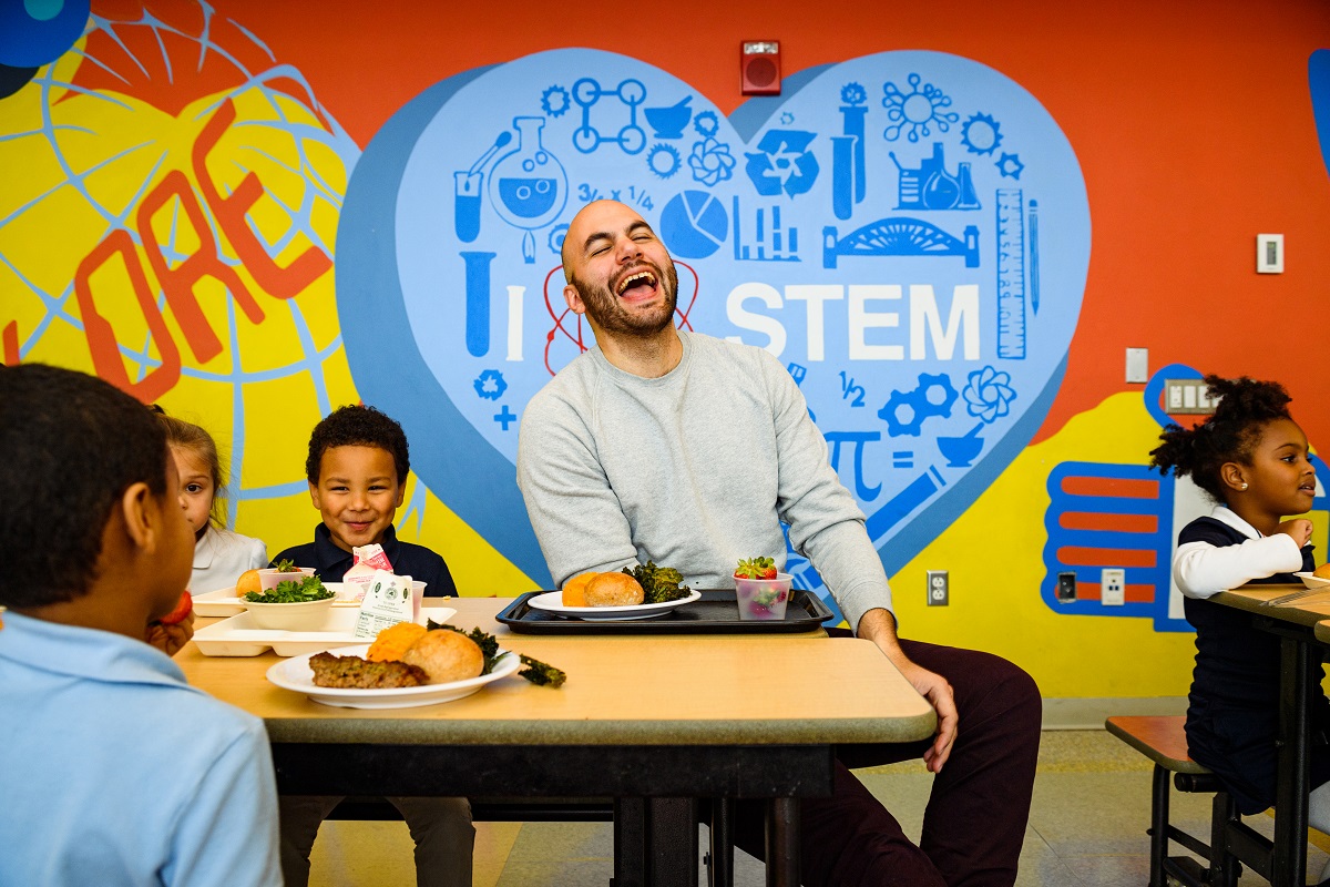 A man, laughing, sits at a table with children, all with trays of food, in front of a brightly-painted wall