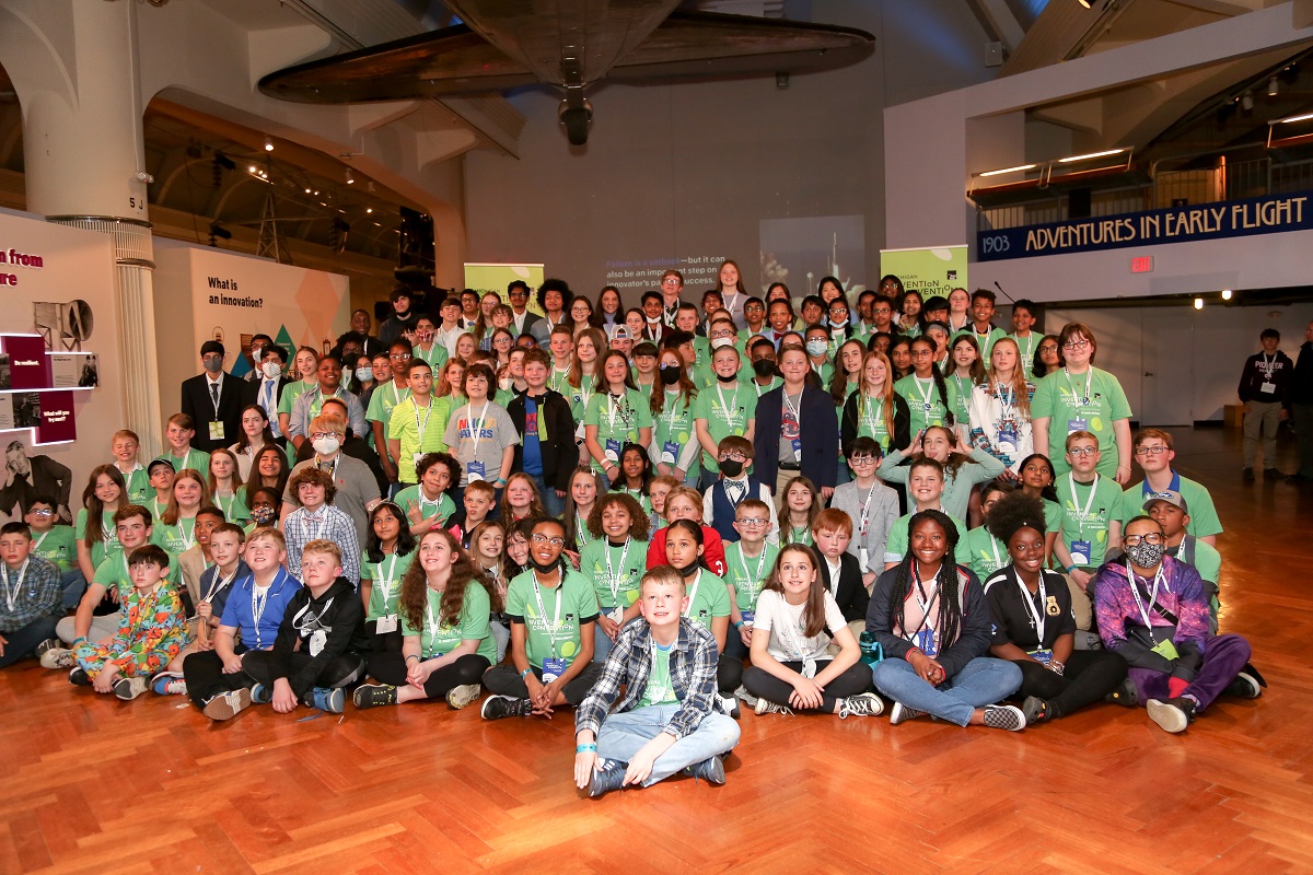 Invention Convention Michigan 2022 competitors A group of children, many wearing green t-shirts and lanyards, pose on the floor of a museum