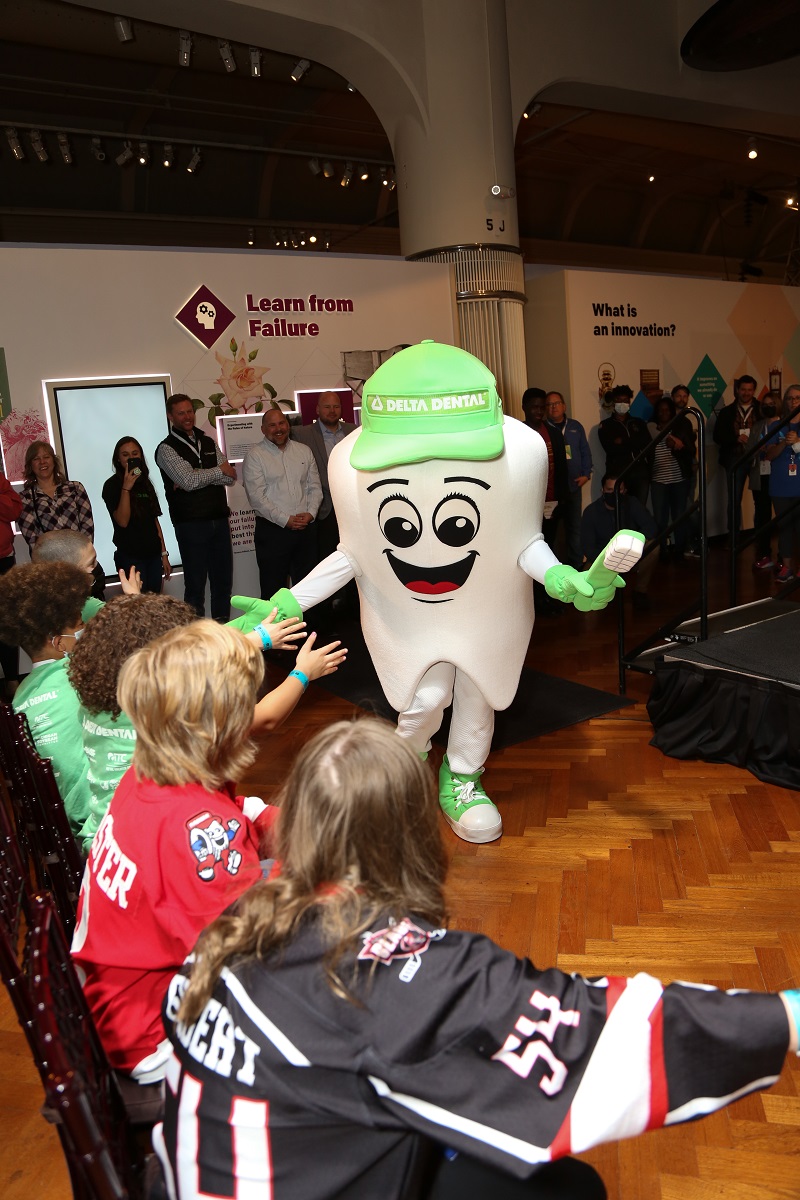 Dazzle from Delta Dental Someone dressed in a tooth costume, with a large smiley face and green hat, gloves, and shoes, entertains a group of people