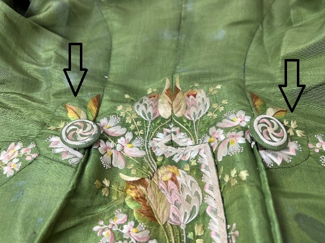 Embroidery on buttons on back of coat Moss-green fabric with elaborate embroidery; arrows point to two buttons covered in embroidered fabric and embroidered semicircles surrounding each