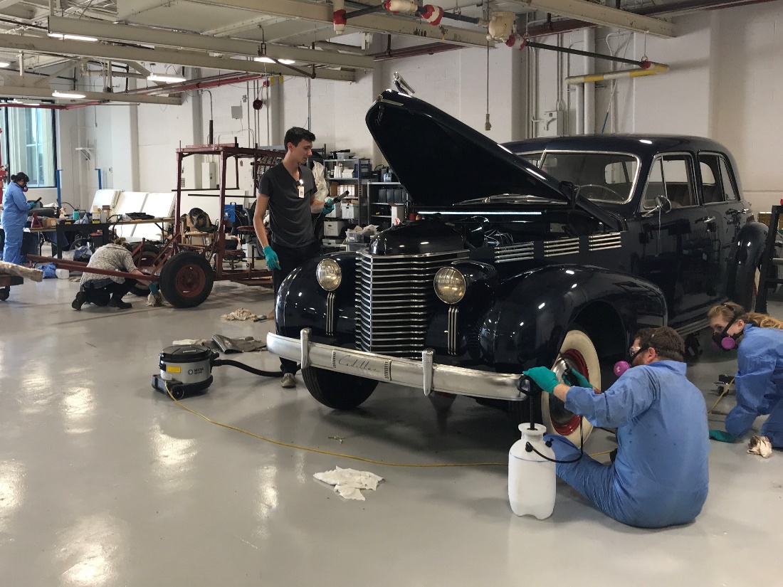 A number of people, some wearing jumpsuits and respirators, work on cars and other equipment in a large room