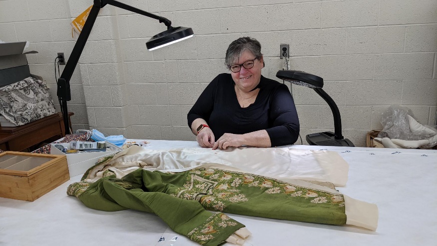 Working on the coat Smiling woman in black glasses and black shirt sits at a work table on which is laid out a moss-green jacket with detailed floral embroidery