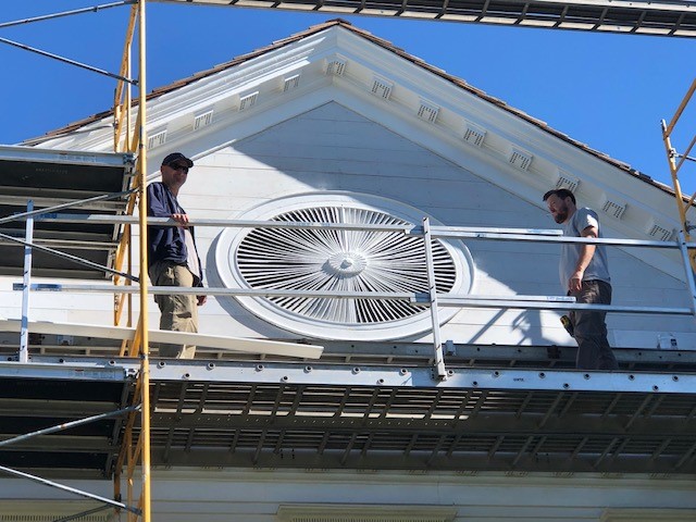 Noah Webster Home ellipse being reinstalled in the pediment Two men on scaffolding stand on either side of a large wooden architectural element on a building façade