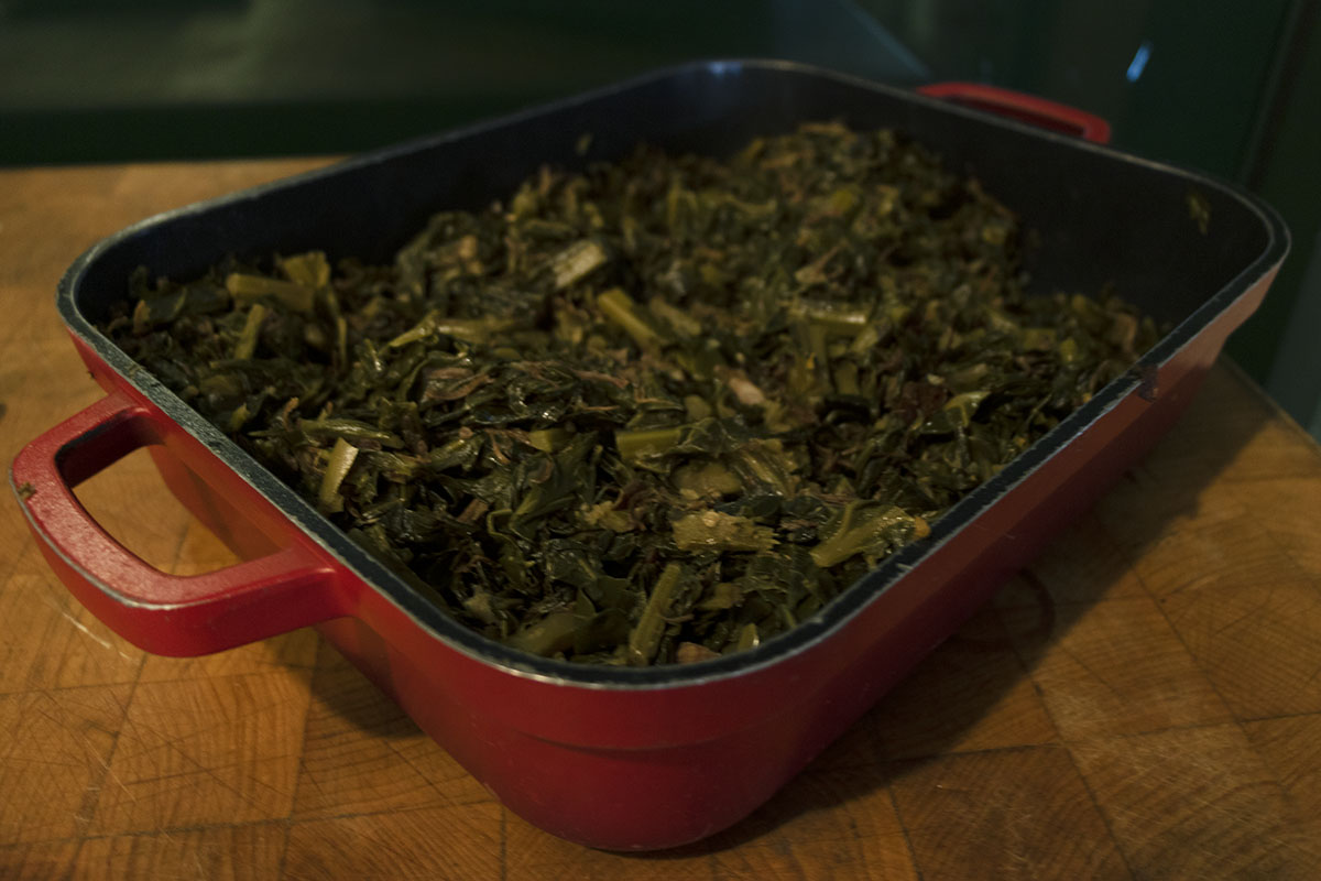 Red casserole dish filled with cooked greens, sitting on butcher block