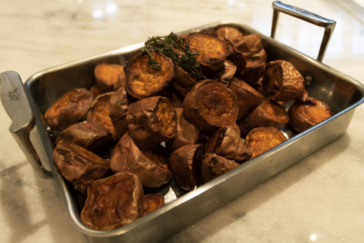 Silver chafing dish containing large chunks of sweet potatoes topped with herbs, sitting on white and gray marble counter
