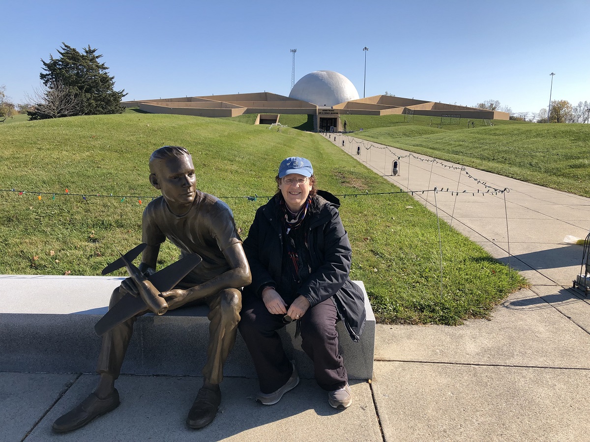Woman sits on bench next to statue of seated person, in front of a large lawn and low building