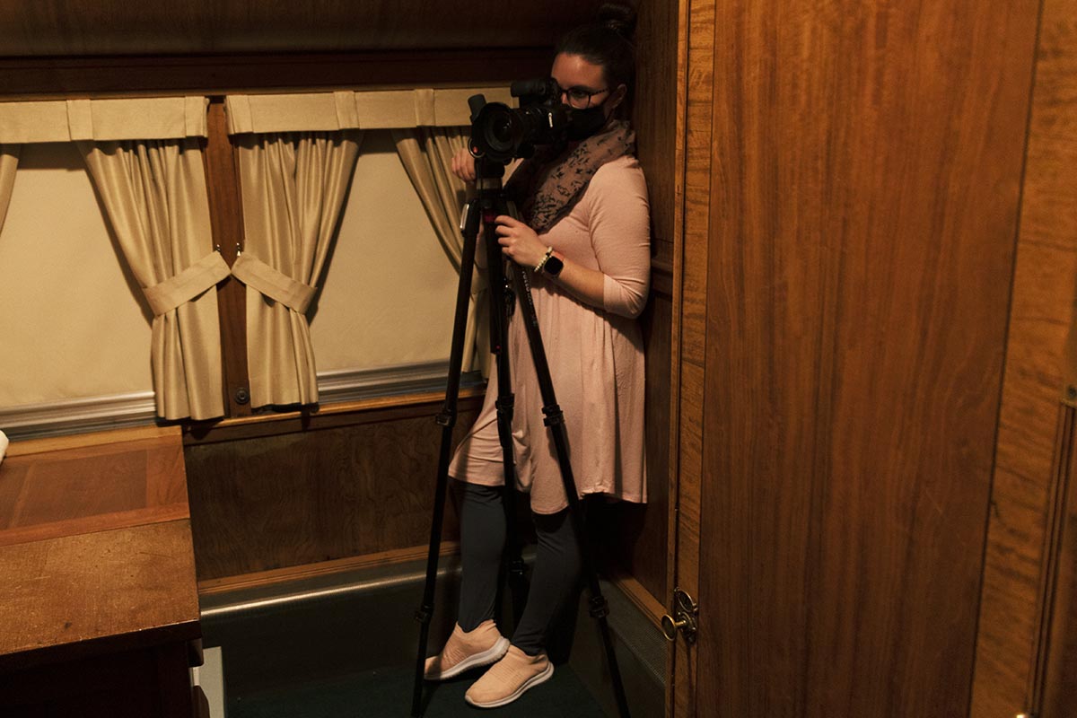 Woman stands in tight corner of wood-paneled room behind a camera on a tripod