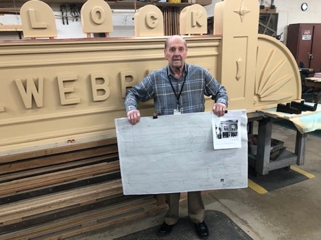 Mose Nowland holds one of his detailed architectural drawings of the C. Webbert sign Man wearing plaid shirt holds drawing in front of large sign in workshop space