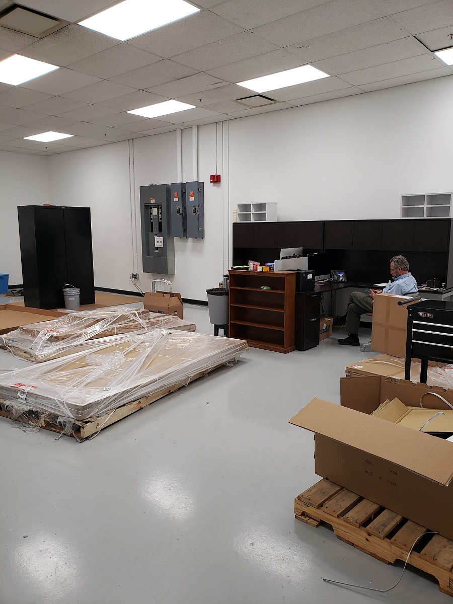 Our new (pre-unpacking) office in the MSB Photo Studio Man sits at empty desk in room containing a few shrinkwrapped piles of wood, pallets, and empty furniture