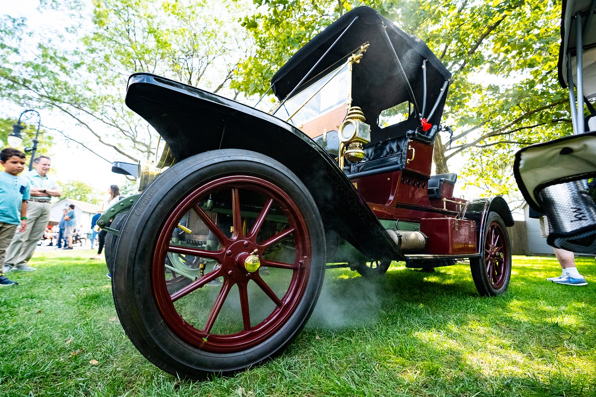 Low-angle photograph of an old-fashioned largely open maroon car exudes steam as spectators look on