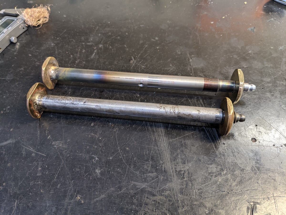 Original and machined hardware Two metal rods with gold stoppers on either end sit on a metal table