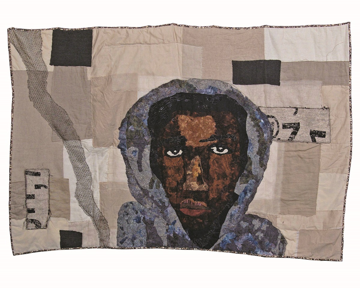 Quilt with background of black and gray blocks of various sizes and face and shoulders of dark-skinned man in a gray hoodie