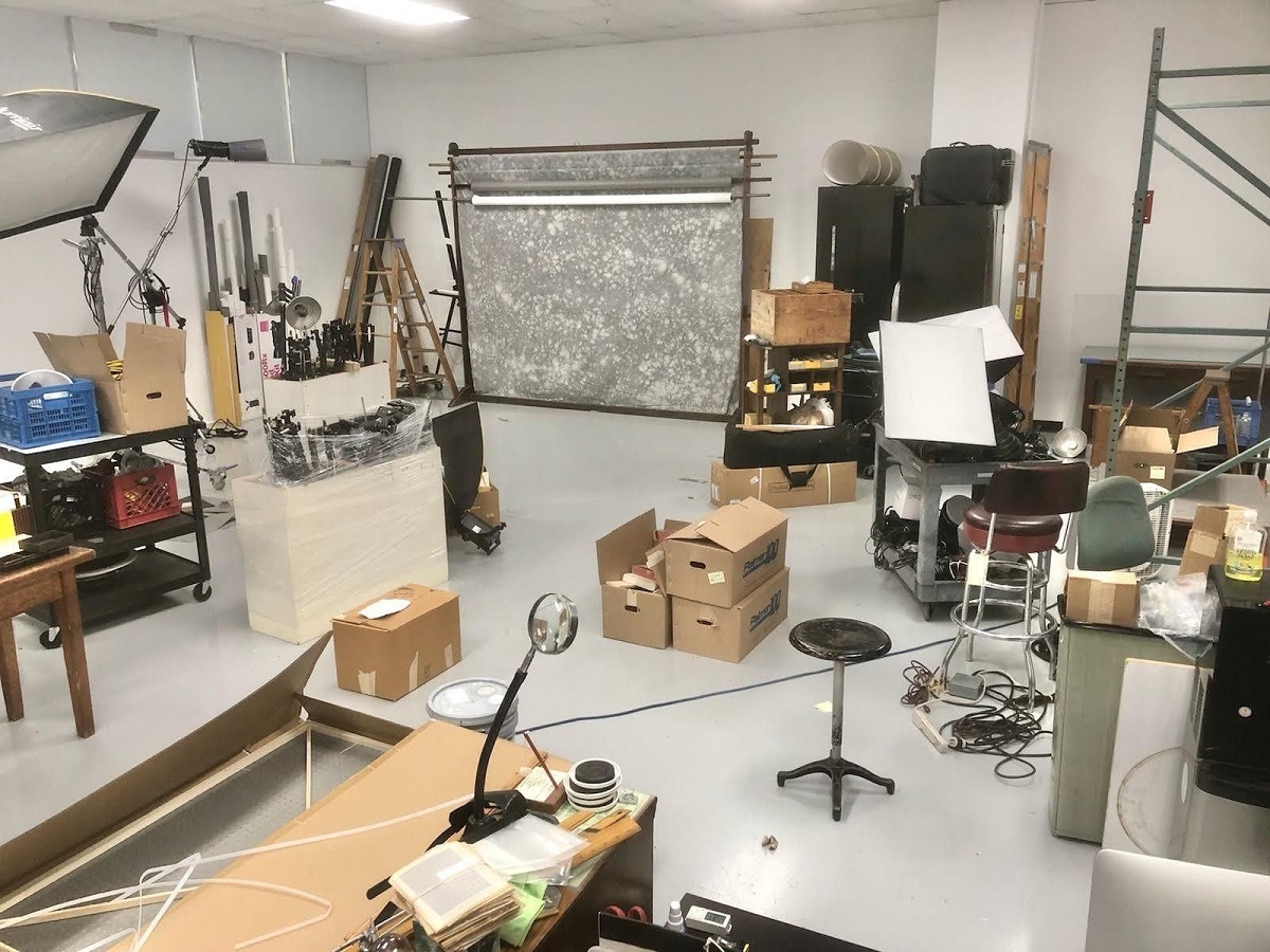 Unpacking and rearranging the new Photo Studio Room cluttered with boxes, furnishings, and other items, with gray and white mottled backdrop in background
