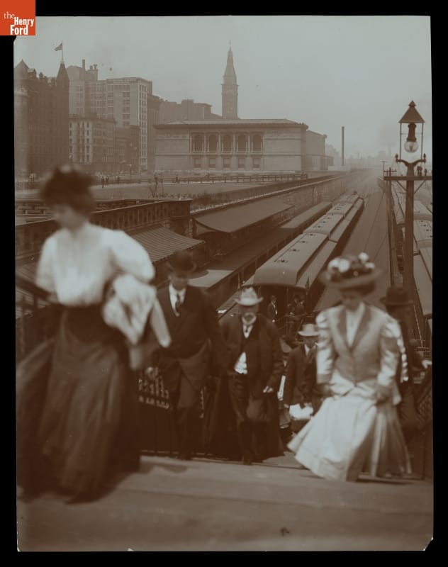People climb a staircase up from sunken railroad tracks with trains on them; buildings in background