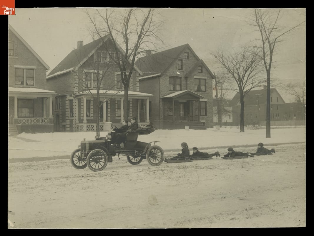Edsel Ford at the Wheel of a Ford Model N with Boys on Sleds in Tow, 1906 Car with two people in it, driving down snowy street with four children on sleds tied to bumper behind; trees and houses in background