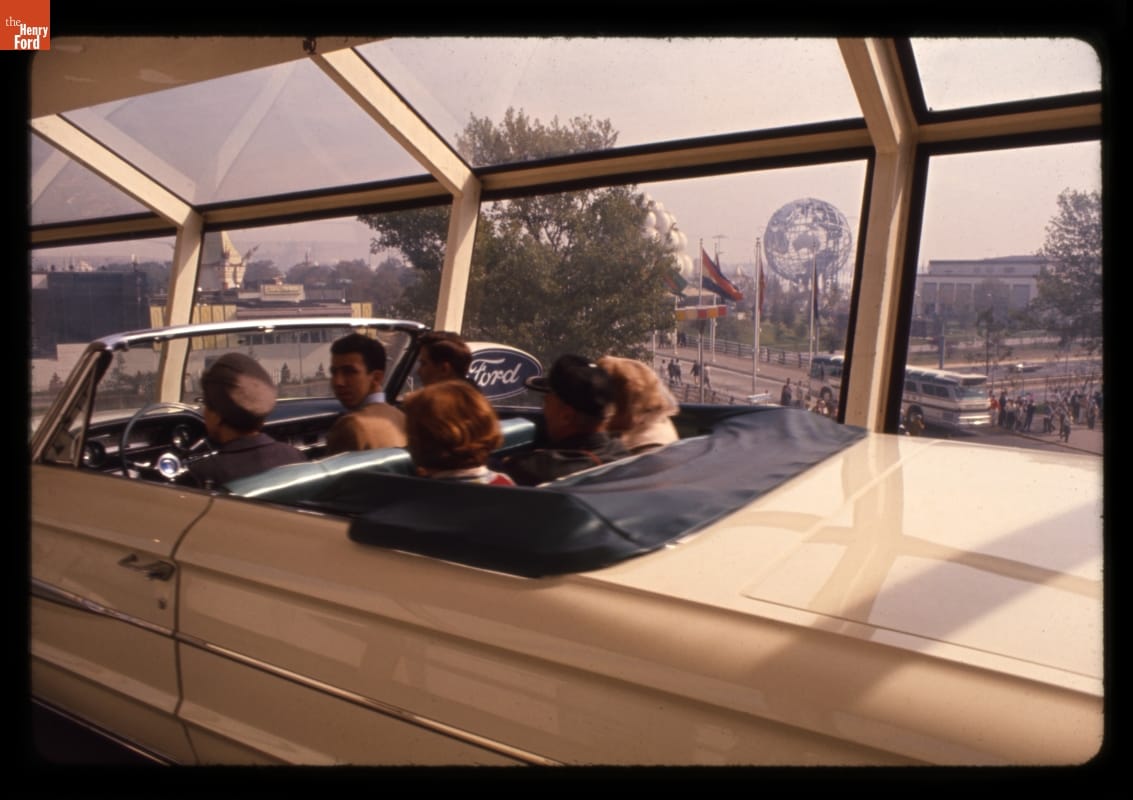 Six people sit in white convertible in a glass tunnel with a fairground visible through the windows