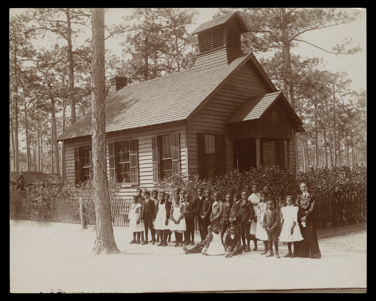 Black-and-white photo of group of African American boys and girls standing with an African American woman outside a small wooden building 