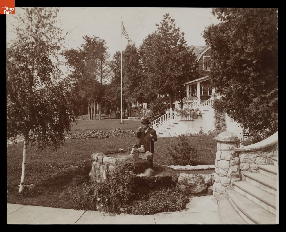 Man Drinking from Flowing Well, Wequetonsing, Michigan, circa 1906 Black-and-white photo of man in suit drinking from stone well on a lawn near houses or other buildings