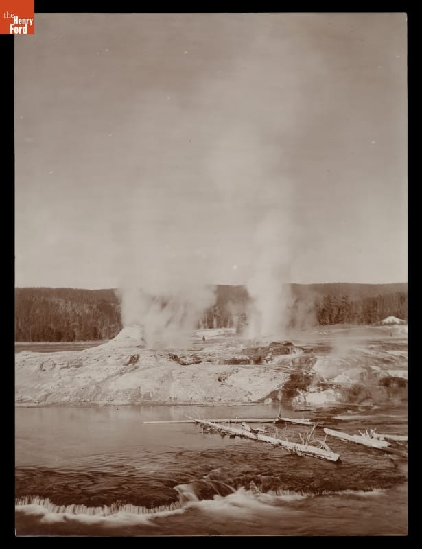 Geysers at Yellowstone National Park, Black-and-white photo of geysers erupting steam and/or water into the air in a rocky landscape with a lake or river in the foreground and forest in the background