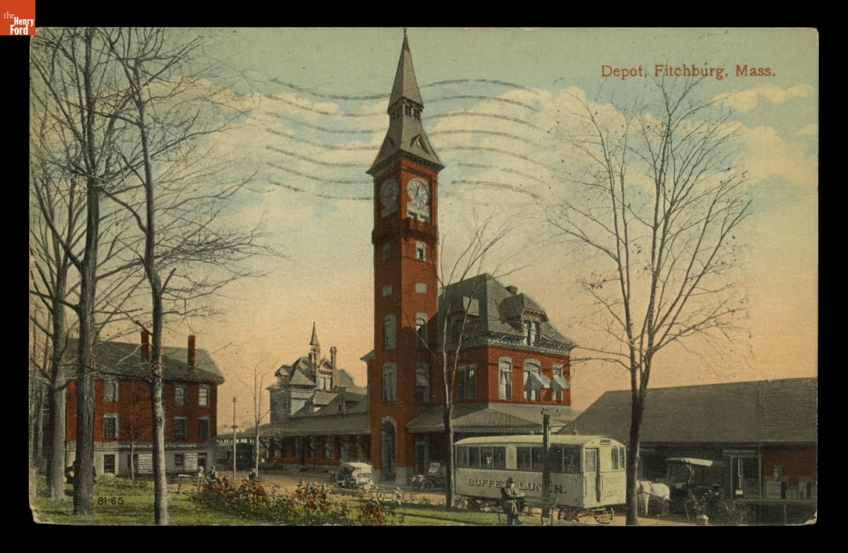 Color postcard of red brick buildings, one with tall clocktower, with cars, wagon, and green space in front
