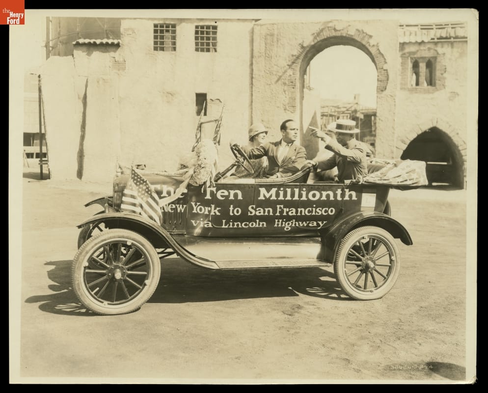 Black-and-white photo of three people sitting in an early open car with text on side, parked in front of buildings and a masonry arch