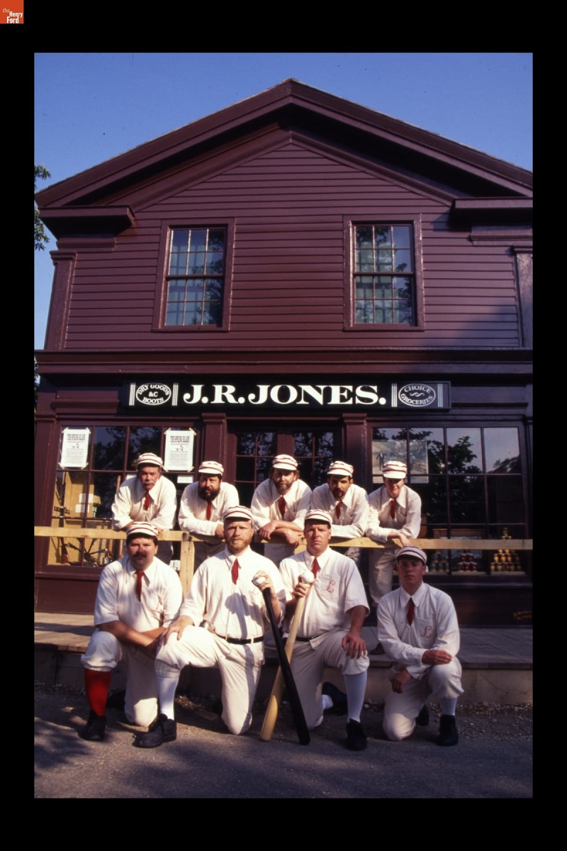 Nine people in old-fashioned baseball uniforms stand and kneel in front of a maroon wooden building with sign