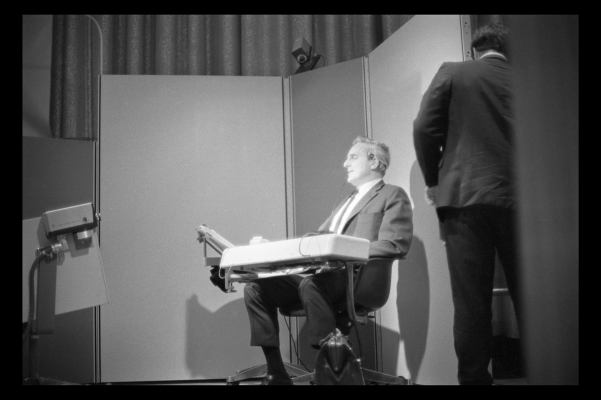 Man sitting in chair with attached console, with video cameras pointed at him; another man to side