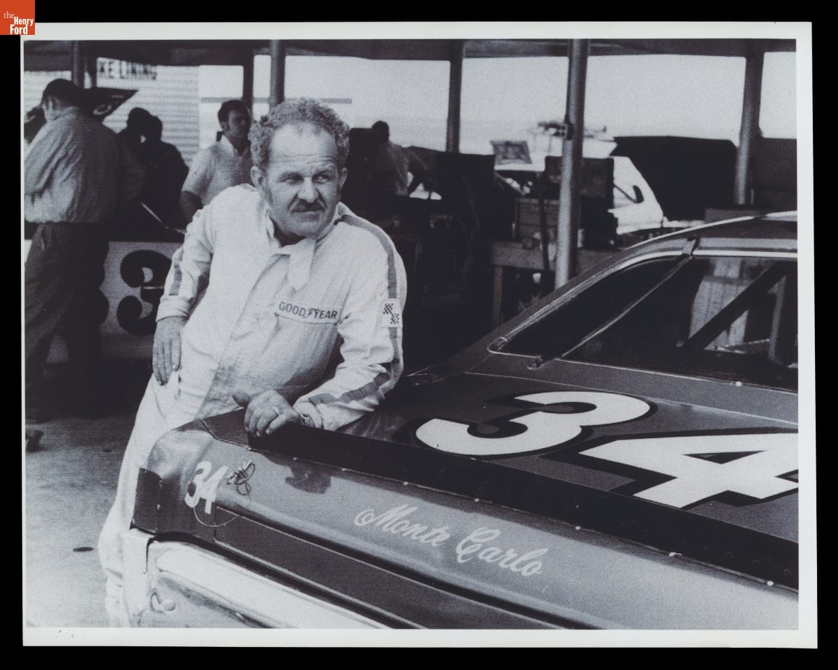 Man in jumpsuit leans against trunk of racecar, with other people and cars in the background