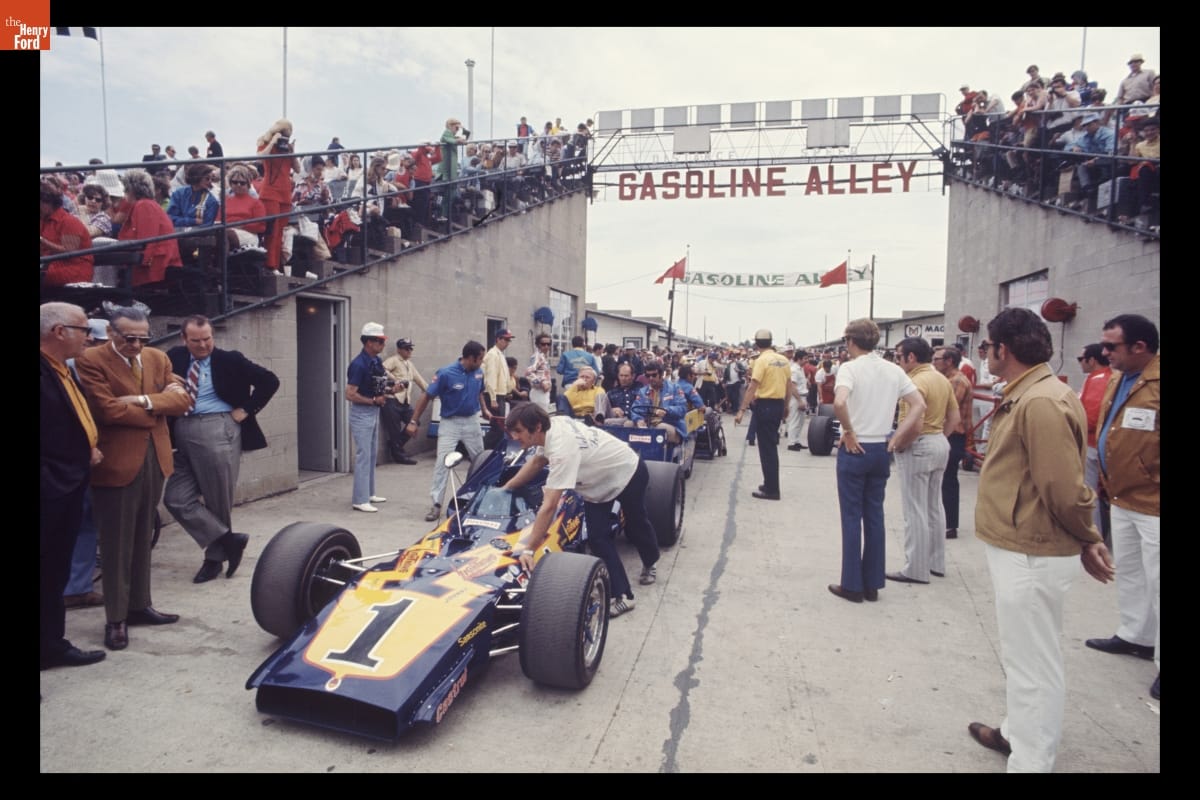 People push racecars through a gap between concrete grandstands filled with people as many watch