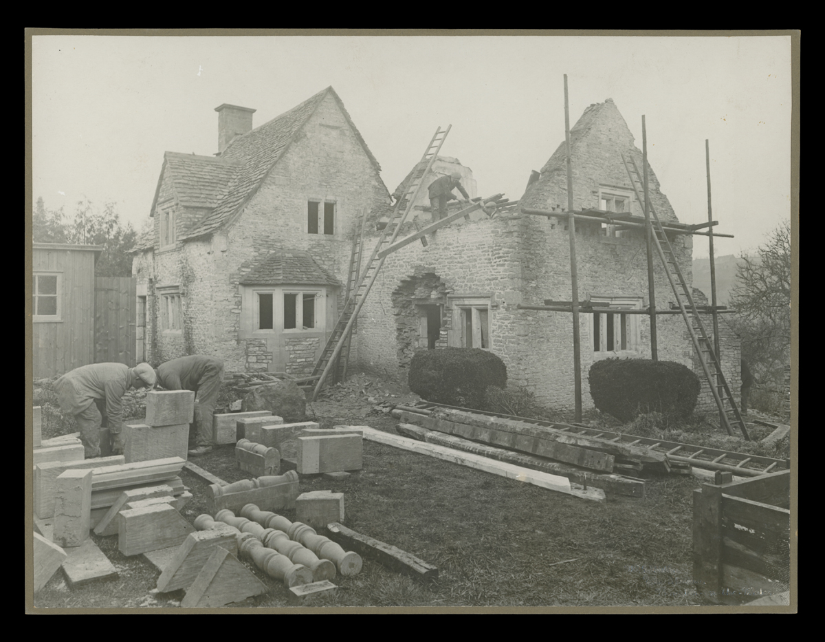 Dismantling Cotswold Cottage at its Original Site in England, 1929-1930 Partially disassembled stone building, with scaffolding around it and architectural elements laid out on the grass surrounding it
