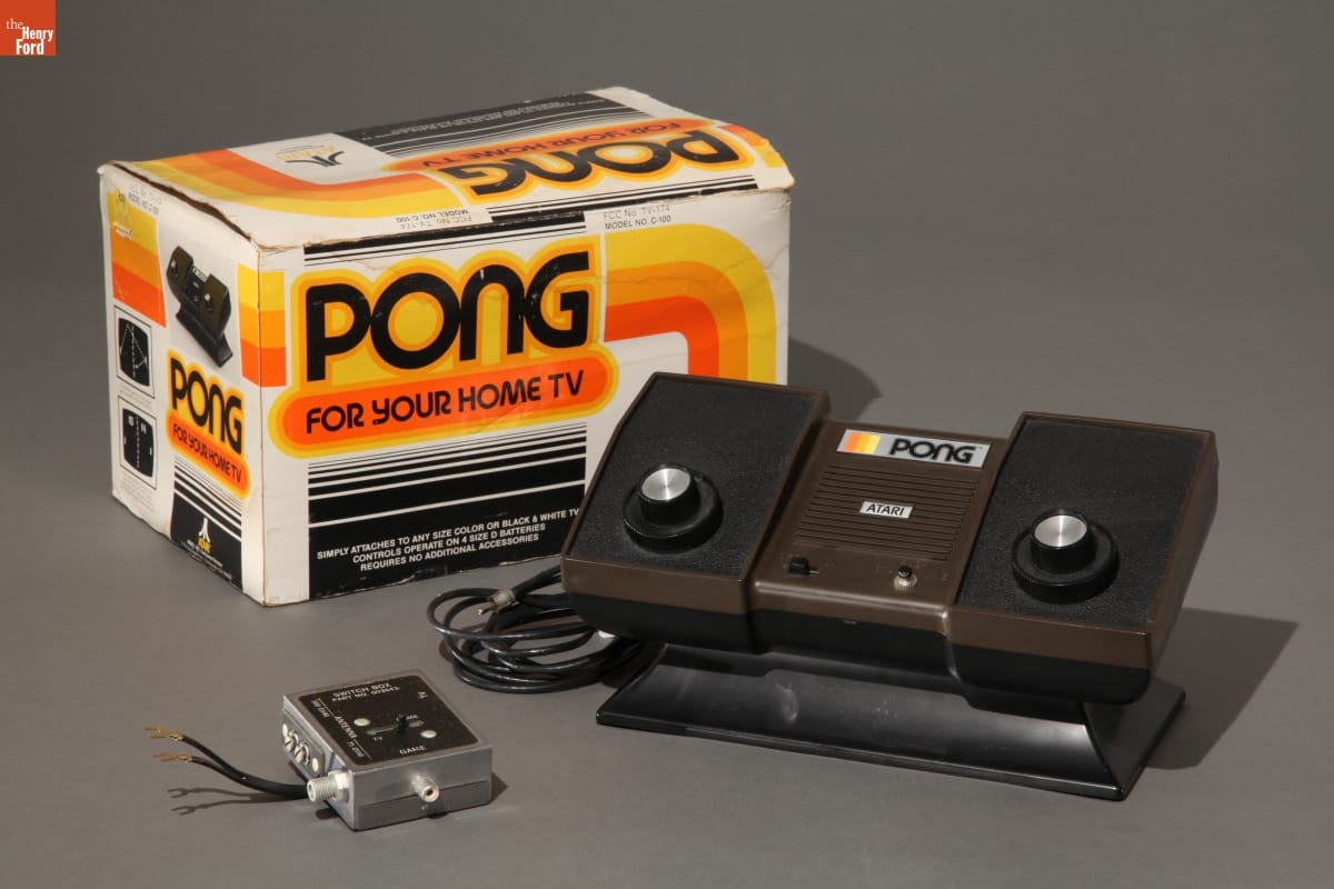 Atari Pong Game, circa 1976 Boxy brown and black video game console with adapter and box with text and graphics in orange, yellow, and black