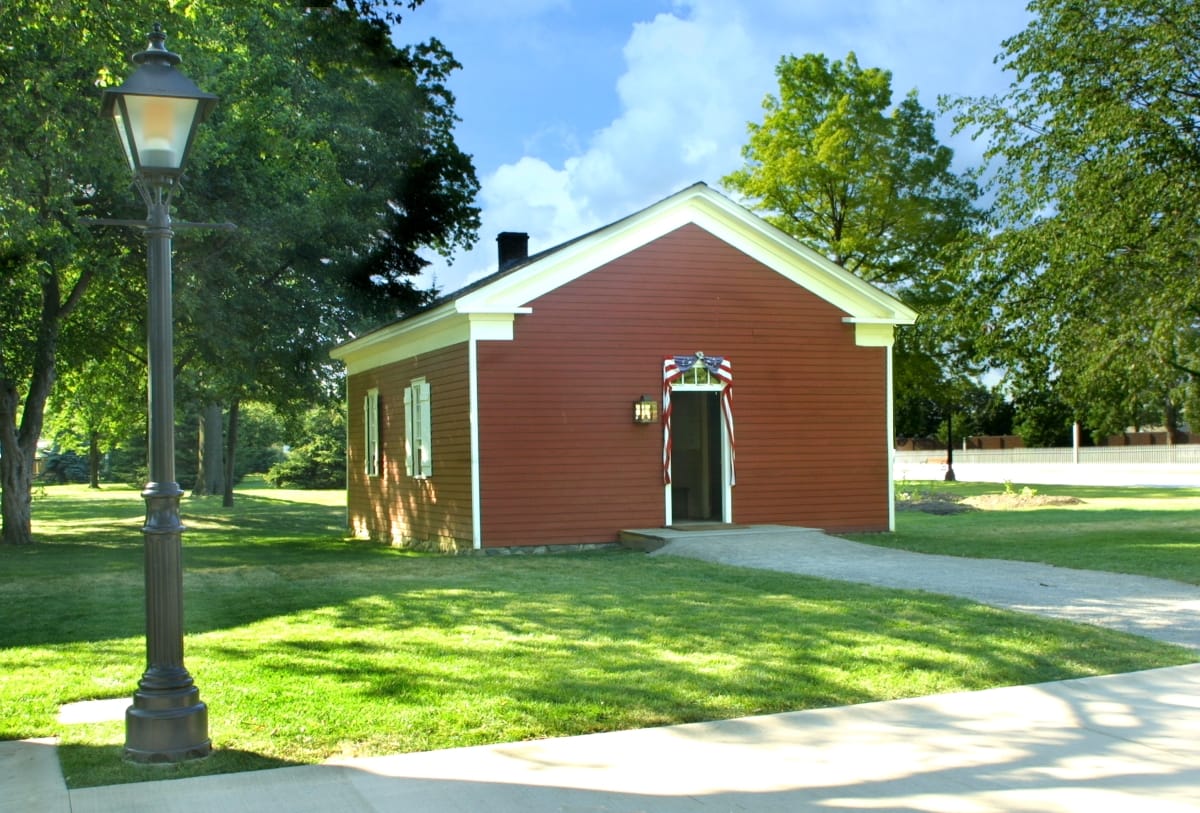 Small red building with white trim set on green lawn with trees, roads, and a streetlamp nearby
