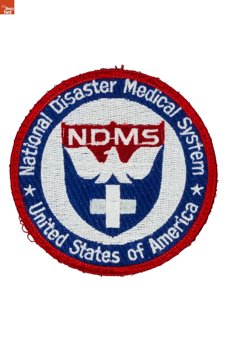 Red, white, and blue round fabric patch with shield containing cross and bird in center; also contains text