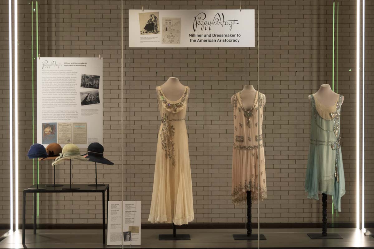 Large glass display case with three dresses, four hats, and signage with text and images