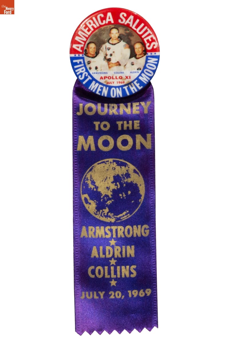 Blue satin ribbon with gold text, topped by medallion with photo of three astronauts inside blue and red semicircles containing text