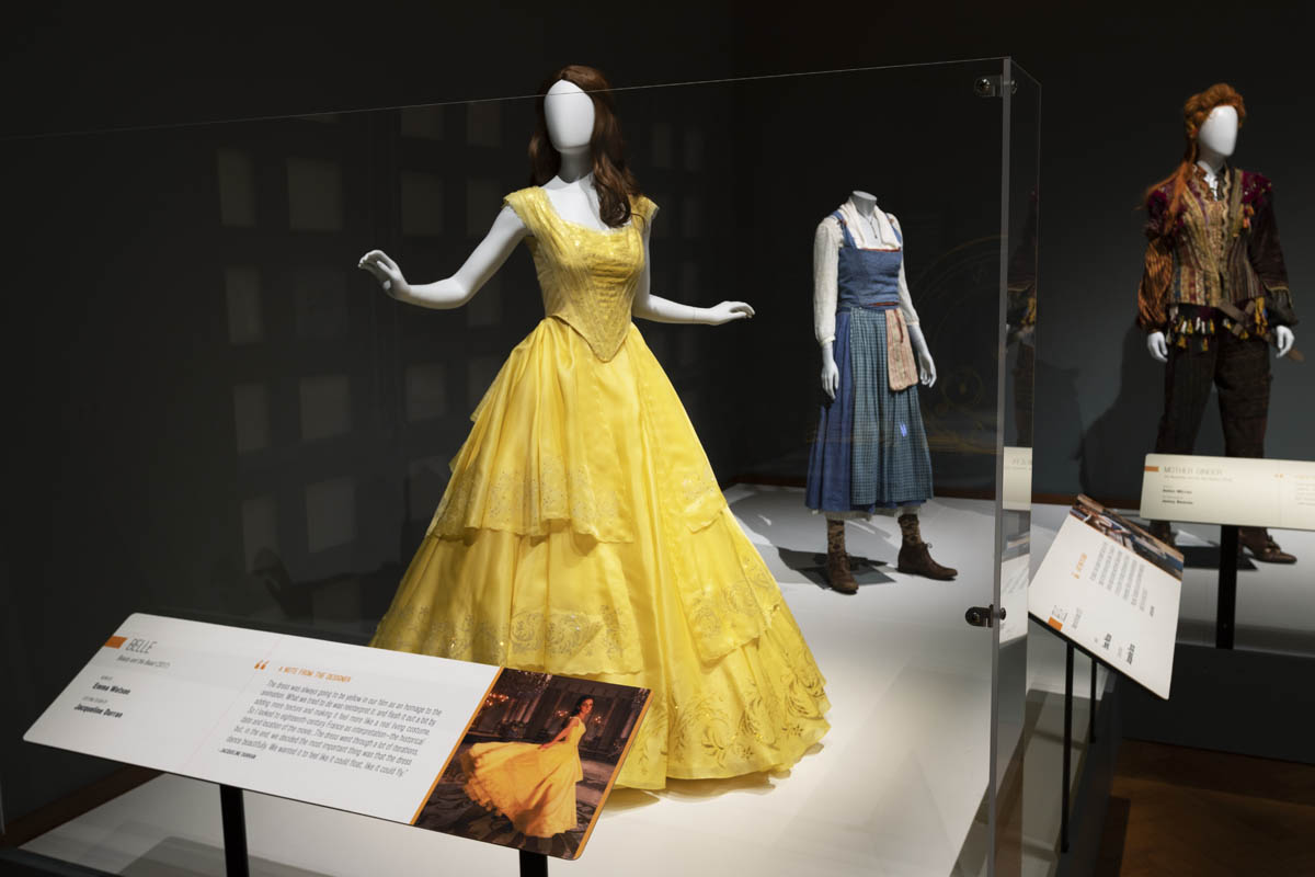 Display case containing mannequins wearing a yellow dress, simple blue jumper, and pants with elaborate jacket; large labels are posted in front of the display