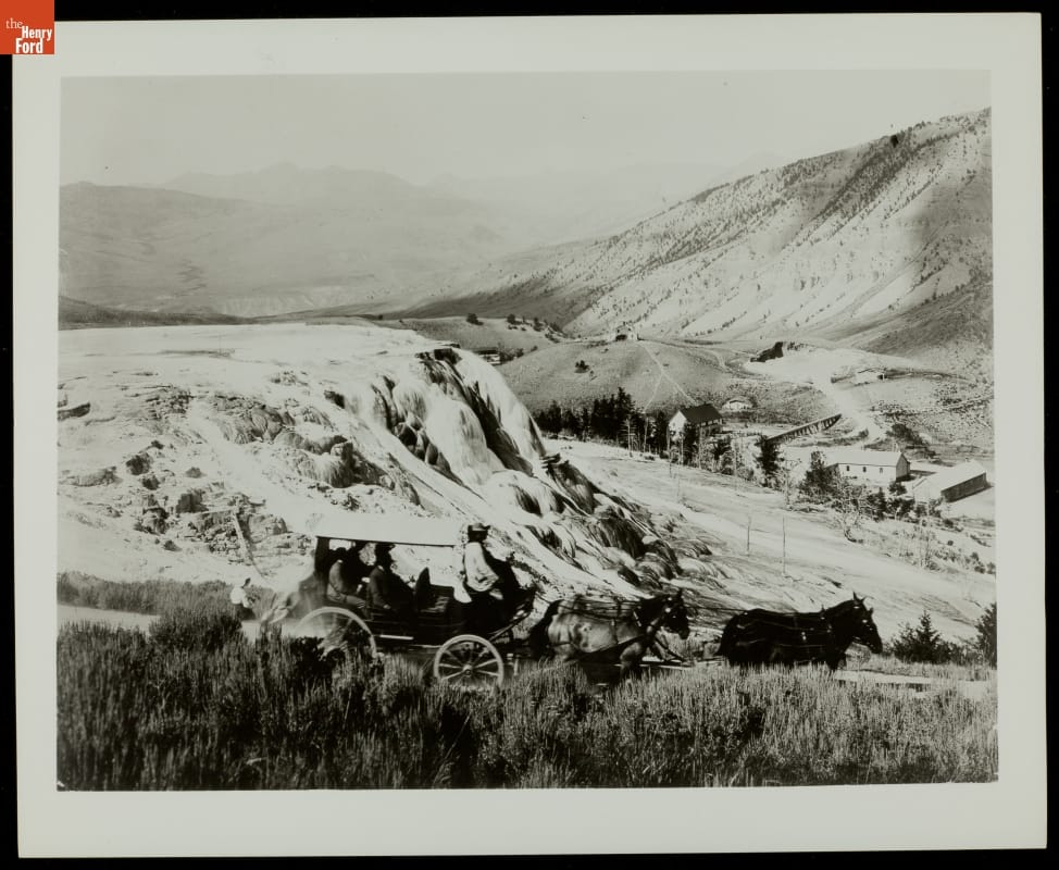 Viewing Jupiter Terrace from a Carriage, Yellowstone National Park, circa 1895 Black-and-white photo of horse-drawn carriage driving past a large rock formation among steep hills and valleys