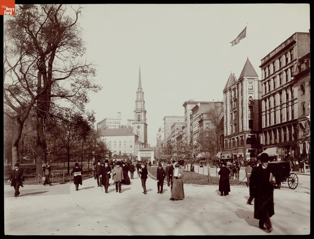 Black-and-white photo of people strolling through a park with a road and buildings visible to the right 