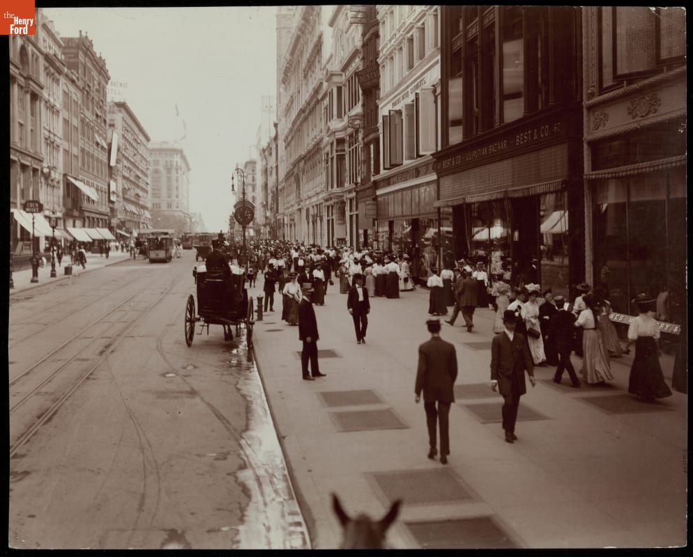 Black-and-white photo of city street with tall buildings and wide sidewalks crowded with people; carriages and streetcars visible on street