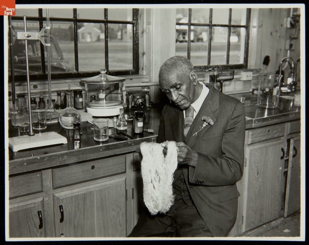George Washington Carver Examining Soy Fiber, Soybean Laboratory at Greenfield Village, 1939 Black man in suit with flower in boutonnière sits by a counter full of scientific equipment and examines yarn or fiber
