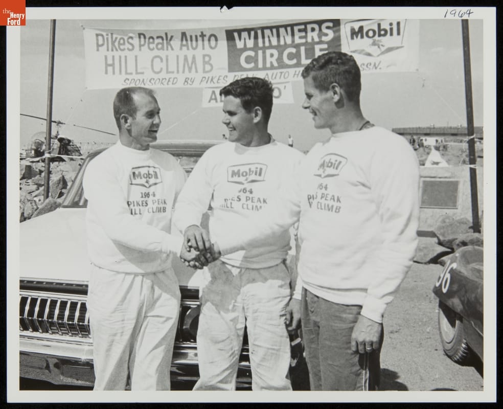 Black-and-white photo of three men in matching sweatshirts joining hands in front of a car and banner