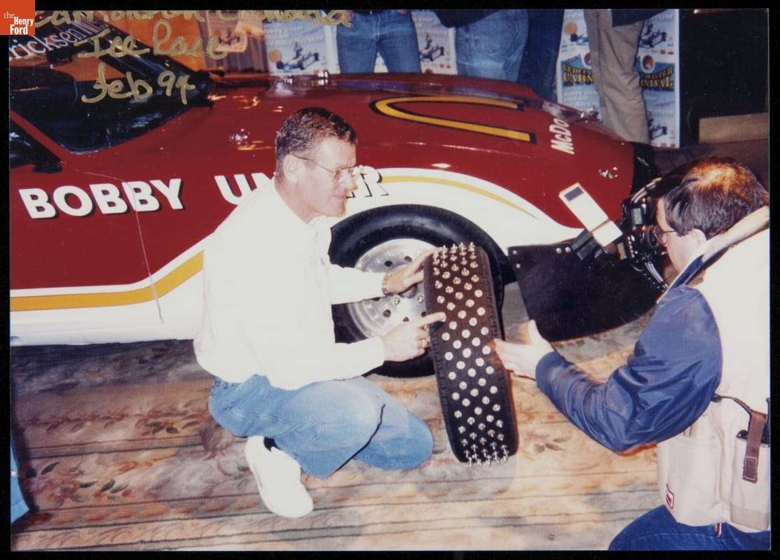 Bobby Unser displaying his tires for ice racing, 1994 Man kneels next to a car pointing to a studded tire he is holding, while another man kneels nearby