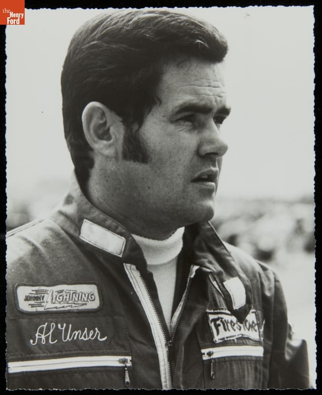 Black-and-white portrait of man with sideburns wearing jacket or jumpsuit with text and logos