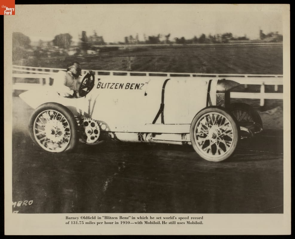Man behind wheel of early open race car on a track; also contains text