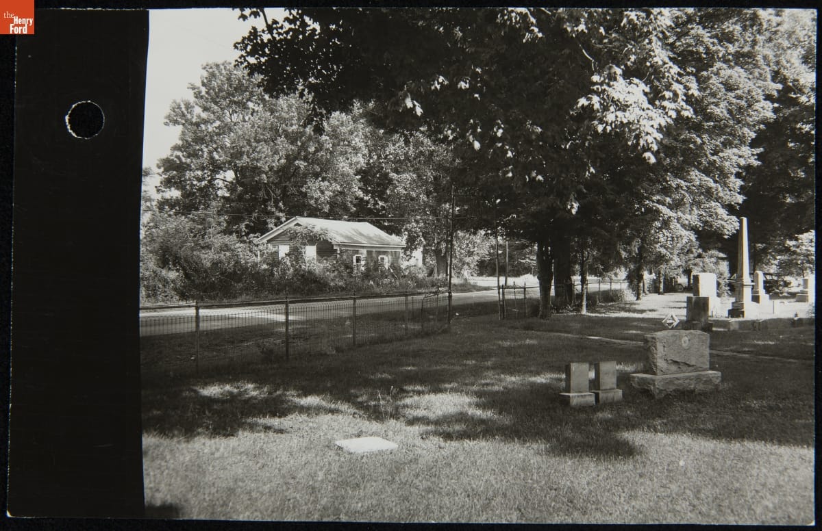 Black-and-white photo of cemetery, road, and small, overgrown building across the road